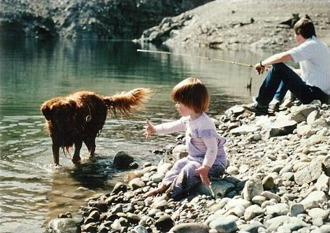 Young Kristine throwing a rock into Lake Koocanusa for her golden retriever while her father fishes